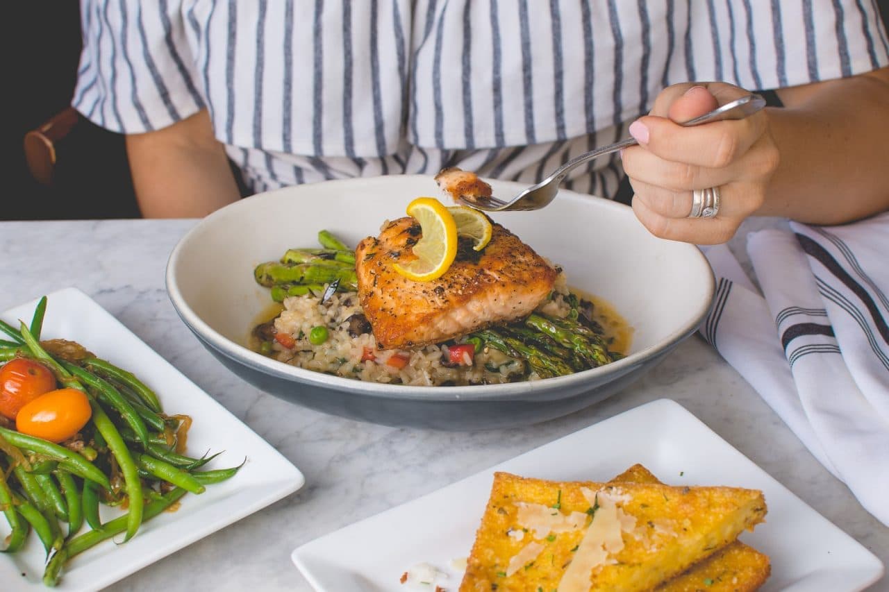 A person in front of a dinner plate of grilled fish and vegetables.