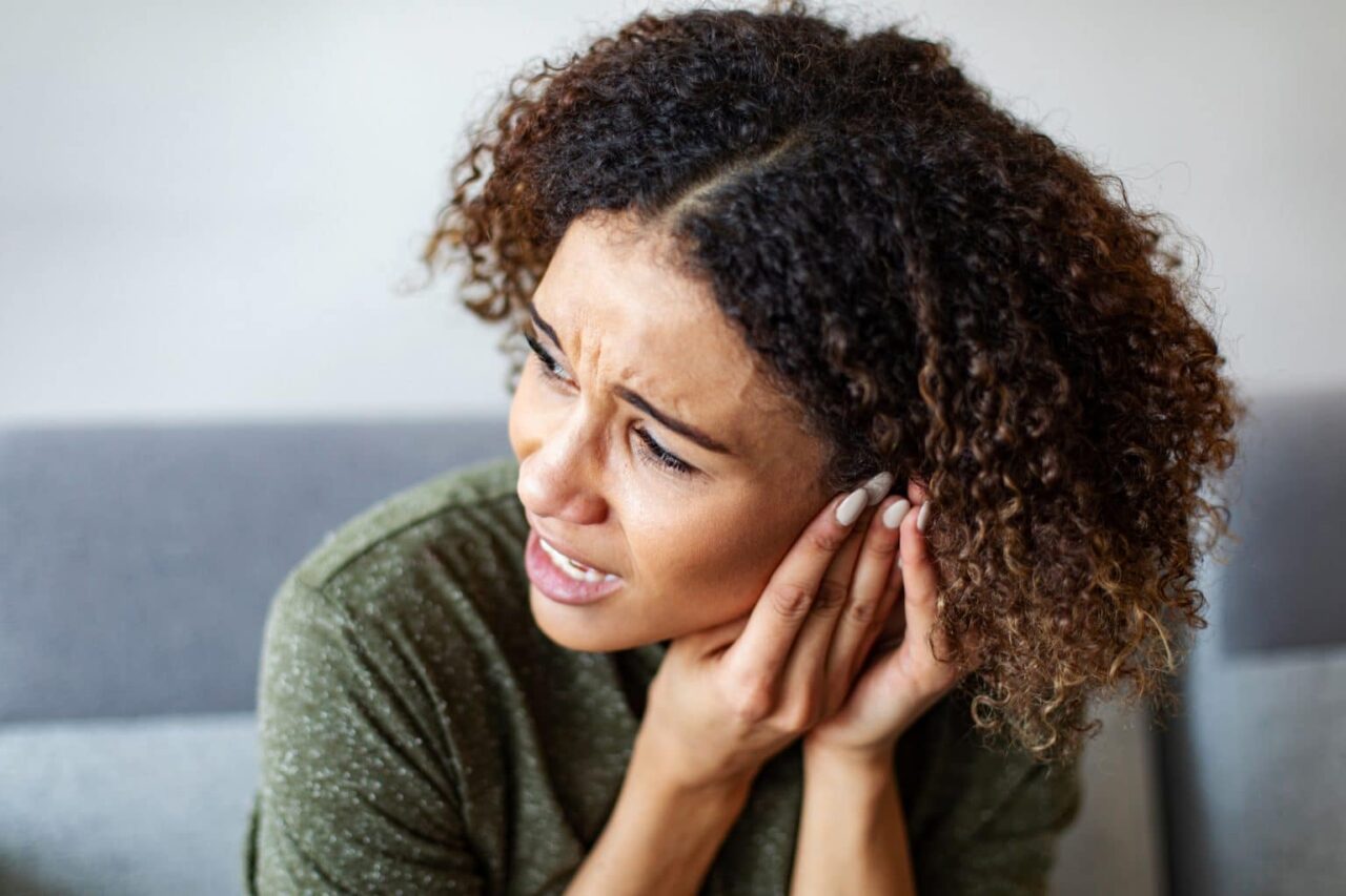 A woman experiencing ear pain holds her hands to her left ear.